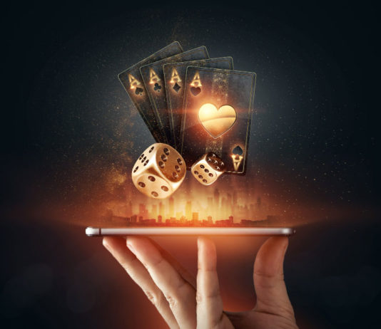 A hand holding a smartphone horizontally, from which glowing golden playing cards and dice are digitally emerging, symbolizing online gambling and casino entertainment. The background is dark with a subtle illuminated cityscape silhouette, enhancing the futuristic and dynamic feel of the image.