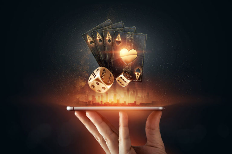 A hand holding a smartphone horizontally, from which glowing golden playing cards and dice are digitally emerging, symbolizing online gambling and casino entertainment. The background is dark with a subtle illuminated cityscape silhouette, enhancing the futuristic and dynamic feel of the image.
